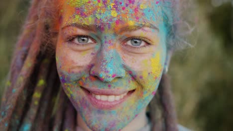 zoom-in portrait of beautiful girl with dreadlocks and colorful face smiling standing outdoors at holi party