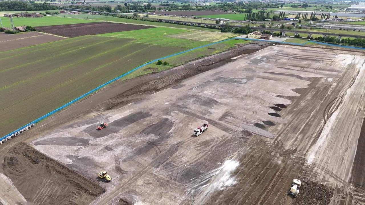 Grandi lavori di sterro per costruzione occupano terreni agricoli coltivati nella Pianura Padana vicino a Fiorenzuola d'Arda, Piacenza, con macchinari pesanti e le Prealpi sullo sfondo, ripresa aerea in allontanamento