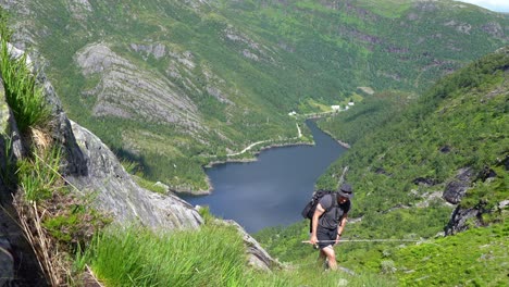 Un-Hombre-Subiendo-Una-Colina-Empinada-Durante-El-Verano-Y-Se-Detiene-Para-Disfrutar-De-Una-Vista-Panorámica-Antes-De-Continuar-Y-Pasar-La-Cámara,-Sosteniendo-Una-Caña-De-Pescar-En-La-Mano-Con-El-Valle-De-Bergsdalen,-Noruega,-Al-Fondo