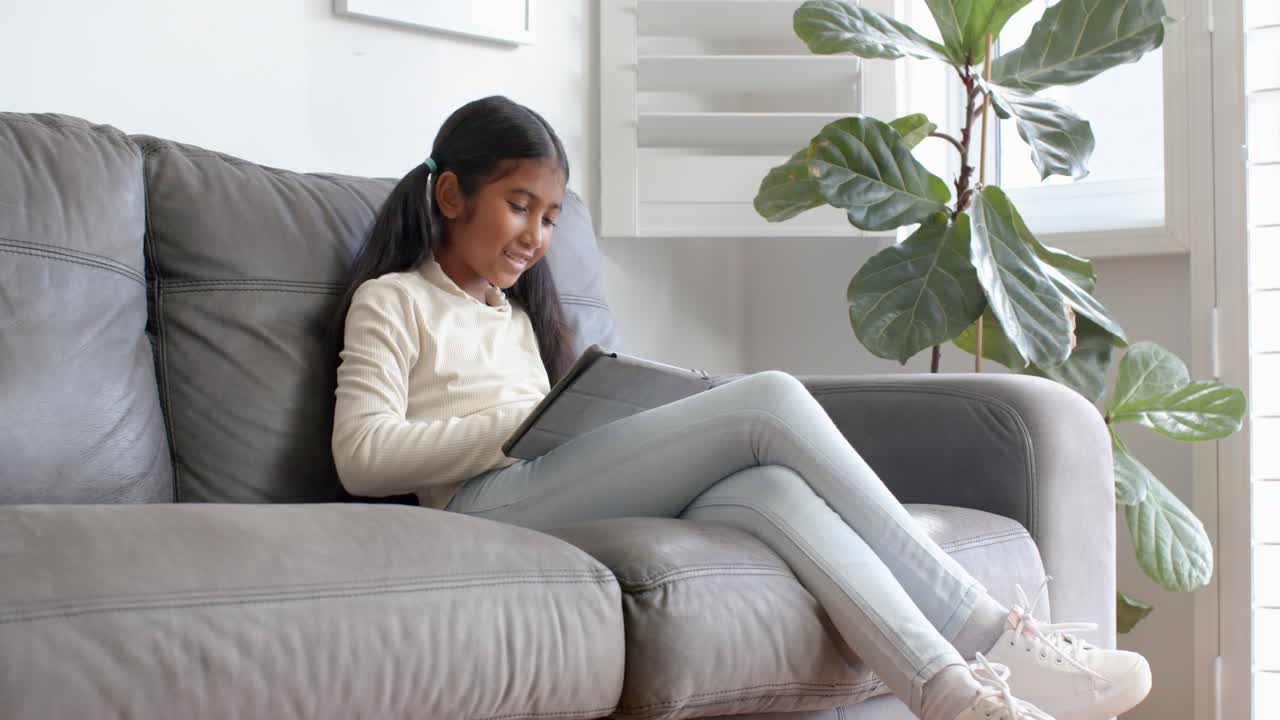 Seduta su un divano grigio, una ragazza asiatica tocca lo schermo del tablet accanto a una pianta di fico a casa per studiare.