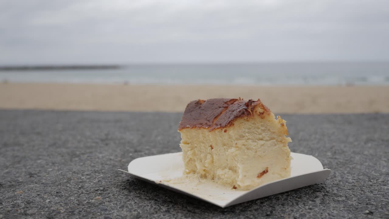 Fresh cheesecake slice on a sandy beach, hand using a wooden spoon to cut