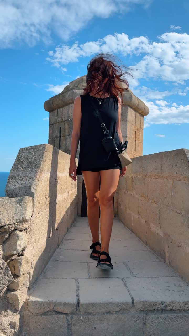 Donna che cammina sulle mura in pietra del Castello di Santa Barbara. Vista dal basso di una turista che cammina lungo le fortificazioni medievali ad Alicante sotto un cielo blu.