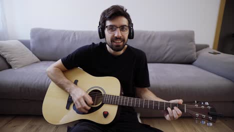 bearded man sitting on the floor, playing on guitar and looking at the camera