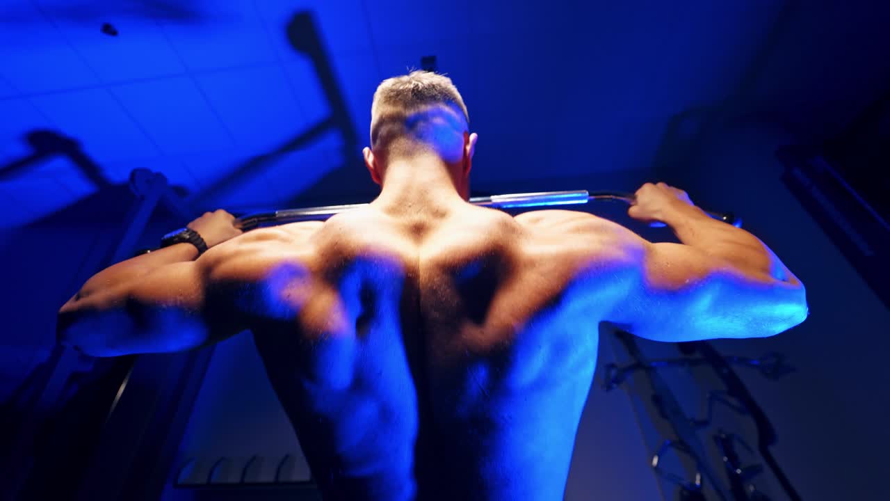 Muscular man tightening on the bar. Backside view of a strong man's body exercising in the gym on blue background. Close-up.