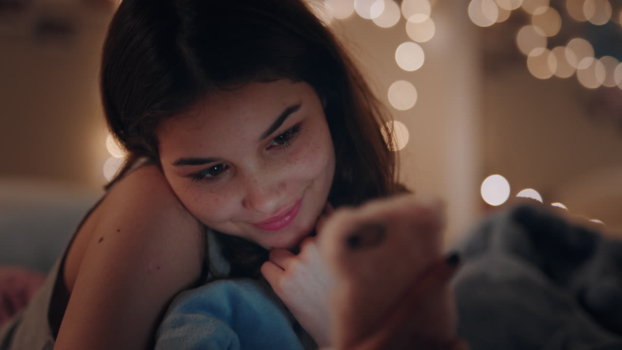 아름다운 십대 소녀가 침대에 누워 스마트폰을 사용하여 문자 메시지를 보내고 소셜 미디어를 검색하고 온라인 채팅을 즐기고 저녁을 집에서 휴식을 취하고 있습니다.