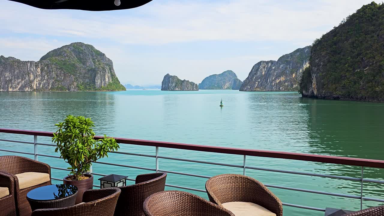 A serene view from a cruise ship's deck restaurant in Ha Long Bay showcases emerald waters surrounded by towering limestone karsts, with comfortable rattan seating and a potted plant in the foreground