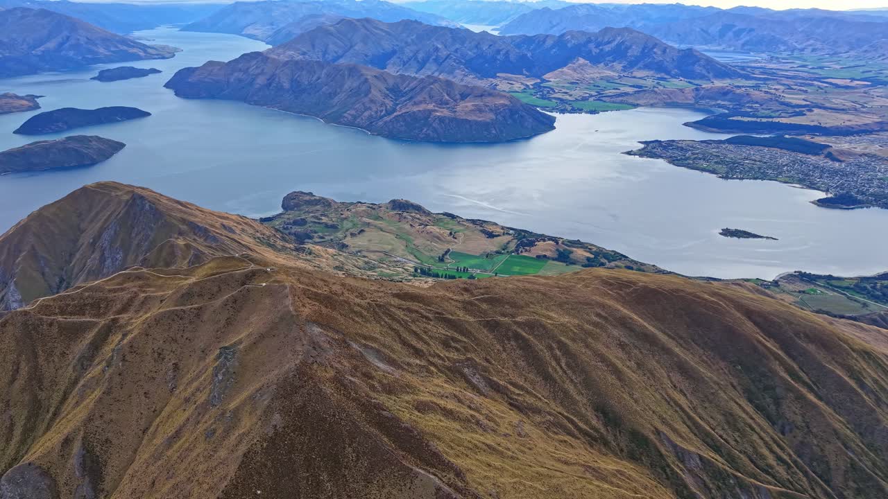 Drone arcs backward above Roys Peak summit, revealing Lake Wanaka, rugged ridges and surrounding Central Otago landscapes under clear daylight
