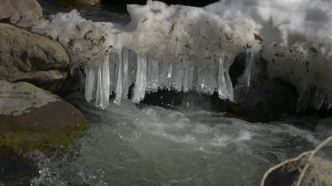 Snow Icicles Over Rocks With A Flowing Stream. Slow Motion, Tilt-down