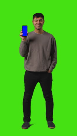 Vertical-Video-Full-Length-Shot-Of-Young-Man-Holding-Blue-Screen-Mobile-Phone-Standing-Against-Green-Screen-1