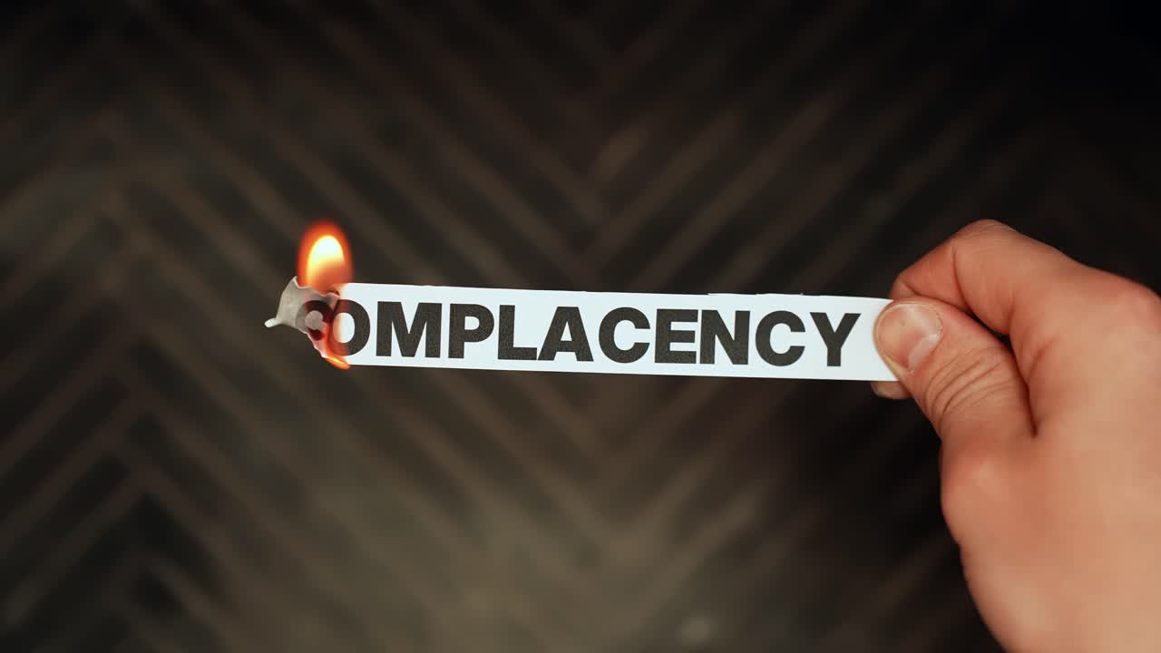 Close-up of a hand holding a paper strip labeled “COMPLACENCY” on fire against a dark blurred background, concept of breaking comfort zone, motivation and change