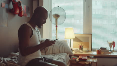 african american man sitting on bed and using smartphone