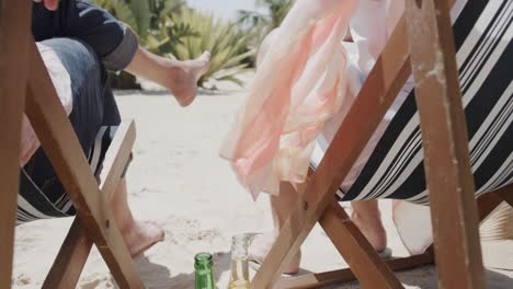 Happy-senior-caucasian-couple-sitting-in-deckchairs-on-beach-reaching-for-beers,-in-slow-motion