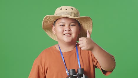 asian tourist boy with a hat and binoculars showing magnifying glass and thumbs up gesture to camera. boy researcher examines something on the green screen background, travel adventure, close up