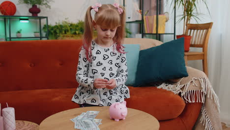 Little-toddler-kid-girl-putting-money-dollar-banknotes-for-future-gift-purchase-at-piggybank-at-home