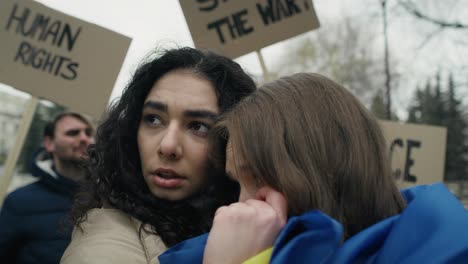 two embracing woman covered with flags and group of young caucasian people manifesting against war in ukraine in the background.