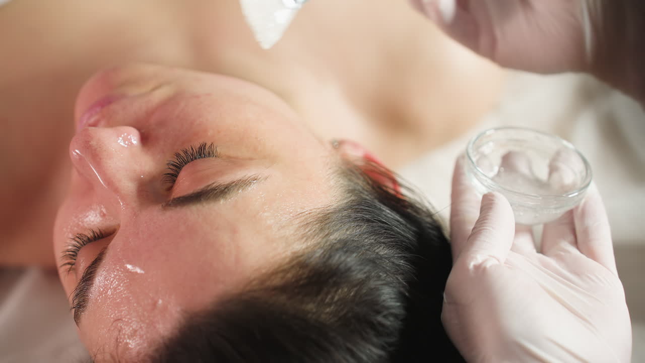 High angle of spa guest lying with eyes closed on bed while skin expert in gloves dips brush into clear gel and applies nourishing facial mask in gentle strokes for glowing radiant skin