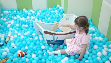 niña en edad preescolar en la sala de juegos seca piscina de remos jugando con pelotas blancas y azules