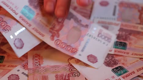 primer plano de una mano contando billetes de cinco milésimas contra un fondo de dinero ruso tendido en una plataforma giratoria.