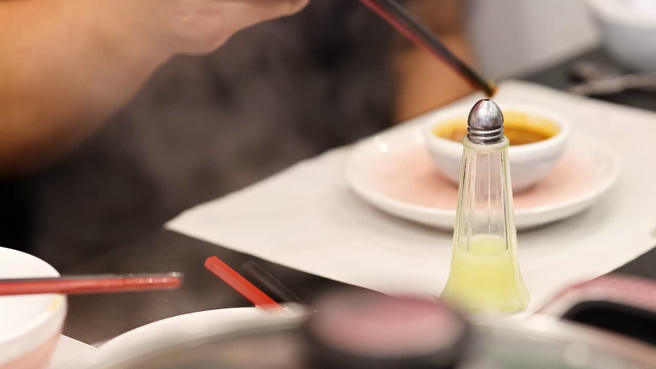 Close-up of hands using chopsticks near a condiment shaker and dipping sauce on a dining table.