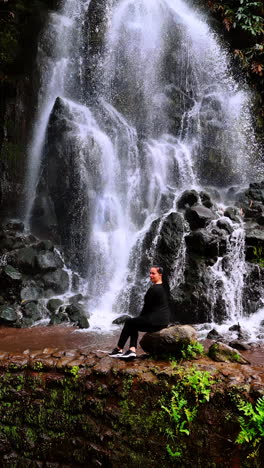 brunette woman sits on the stone looking at beautiful waterfall. water splashes by the black boulders. nature of the azorean islands. vertical video.