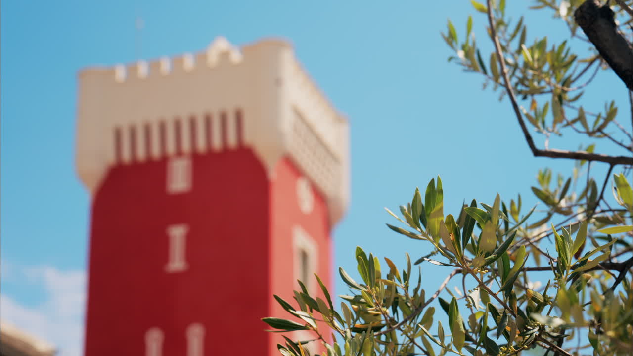 Green tree moving in the wind near the red tower of the Cremat Castle Winery over blue sky