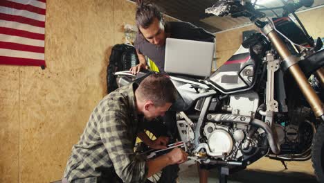 Two-mechanic-guys-are-disassembling-and-inspecting-a-motorcycle.-A-guy-in-a-gray-T-shirt-looks-at-a-laptop-screen-in-a-workshop-with-tools.-Service-center-for-equipment