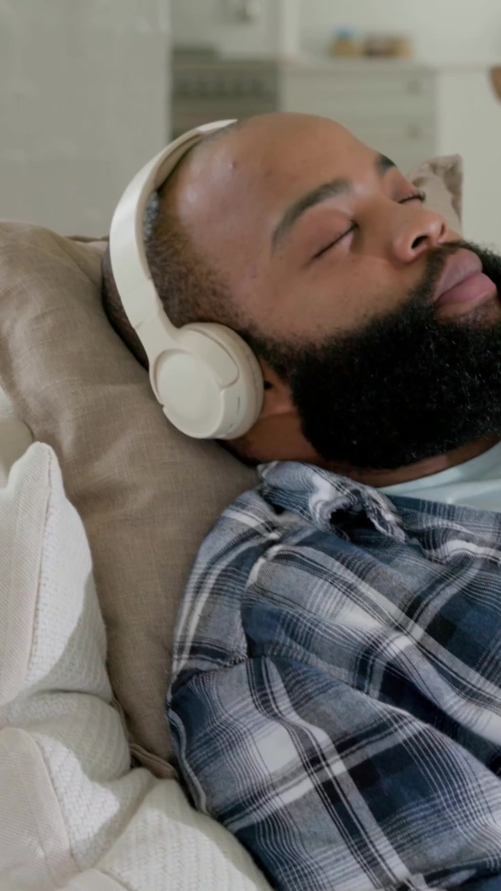 Vertical video: Panning to man reclining and resting on couch at home in plaid shirt with headset