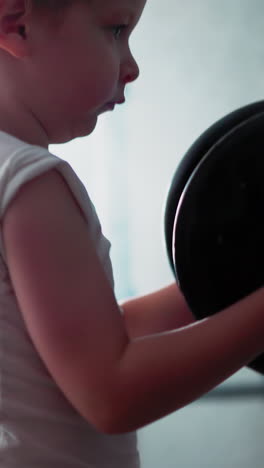 strong little boy lifts heavy barbell standing in gym filled with fake smoke. concentrated toddler does exercise at bright light in gym closeup slow motion
