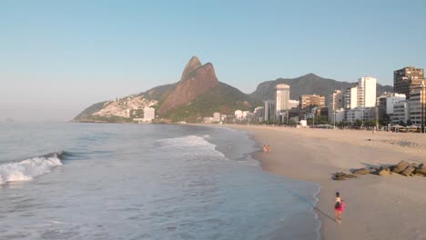 sideways aerial movement accompanying a wave coming in on coastal city beach of rio de janeiro during early morning golden hour seen from above the ocean