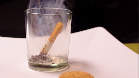 empty-glass-with-tiny-bit-of-ash-placed-on-white-paper-with-black-background-close-up-person-male-hand-throwing-leftover-burning-cigarette-inside-the-cup-while-its-still-on-fire-home-smoking-stress