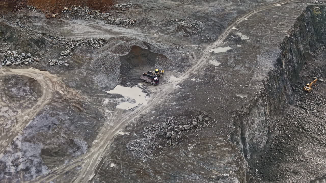 Quarry workers operate heavy machinery at a granite quarry site during daylight in a rugged landscape with piles of stone and earth