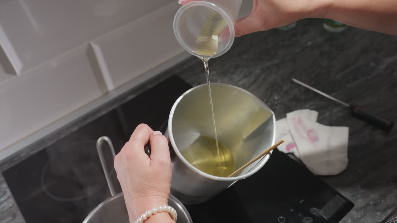 Creative woman pours remaining melted sugar into metal bowl while preparing homemade mixture on kitchen stove with pot of hot water nearby and napkin resting on counter beside tools