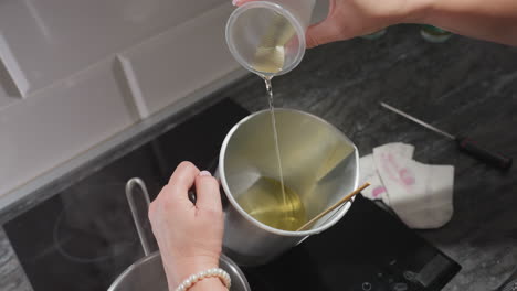 creative woman pours remaining melted sugar into metal bowl while preparing homemade mixture on kitchen stove with pot of hot water nearby and napkin resting on counter beside tools
