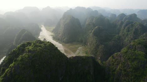 Aerial-Flying-Above-Striking-Limestone-Karsts-Revealing-Ngo-Dong-River-Waving-Between-Mountain-Ranges-and-Line-of-Sampan-Tour-Boats-Traveling-on-Current-in-Ninh-Binh-Vietnam,-Sunset