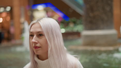 elegant young woman walking toward indoor fountain seating in modern shopping mall under bright ceiling lights surrounded by glass railings polished floor ambient shoppers passing by