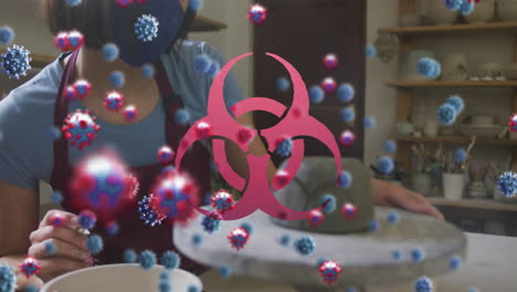 animation of covid 19 cells and biohazard sign over woman working in pottery studio in face mask