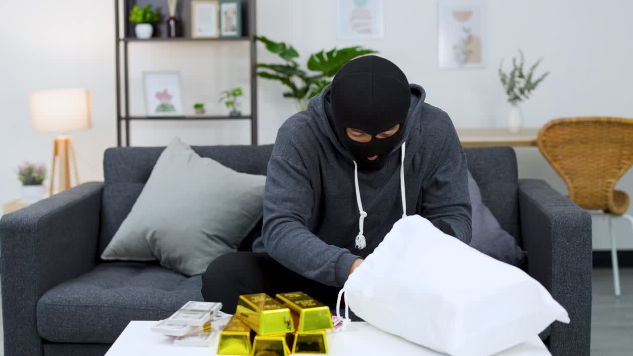 A burglar in a ski mask counts hundred dollar bills beside gold bars and loot