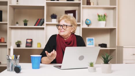 older woman in glasses is talking via video call while sitting at a table with a laptop