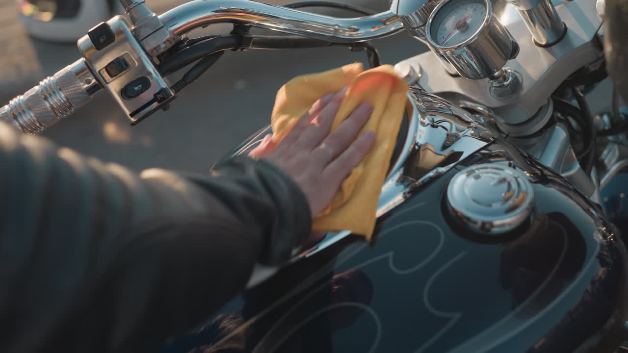 Close up of biker hand in leather jacket cleaning motorcycle tank and handle with yellow cloth, chrome parts reflecting sunlight, focus on motorcycle care and detailed maintenance