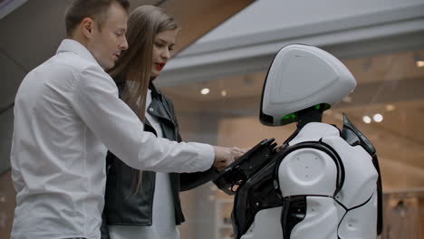 a man and a woman in the mall interact with a robot consultant by tapping the screen and smiling. cyborg android helps people