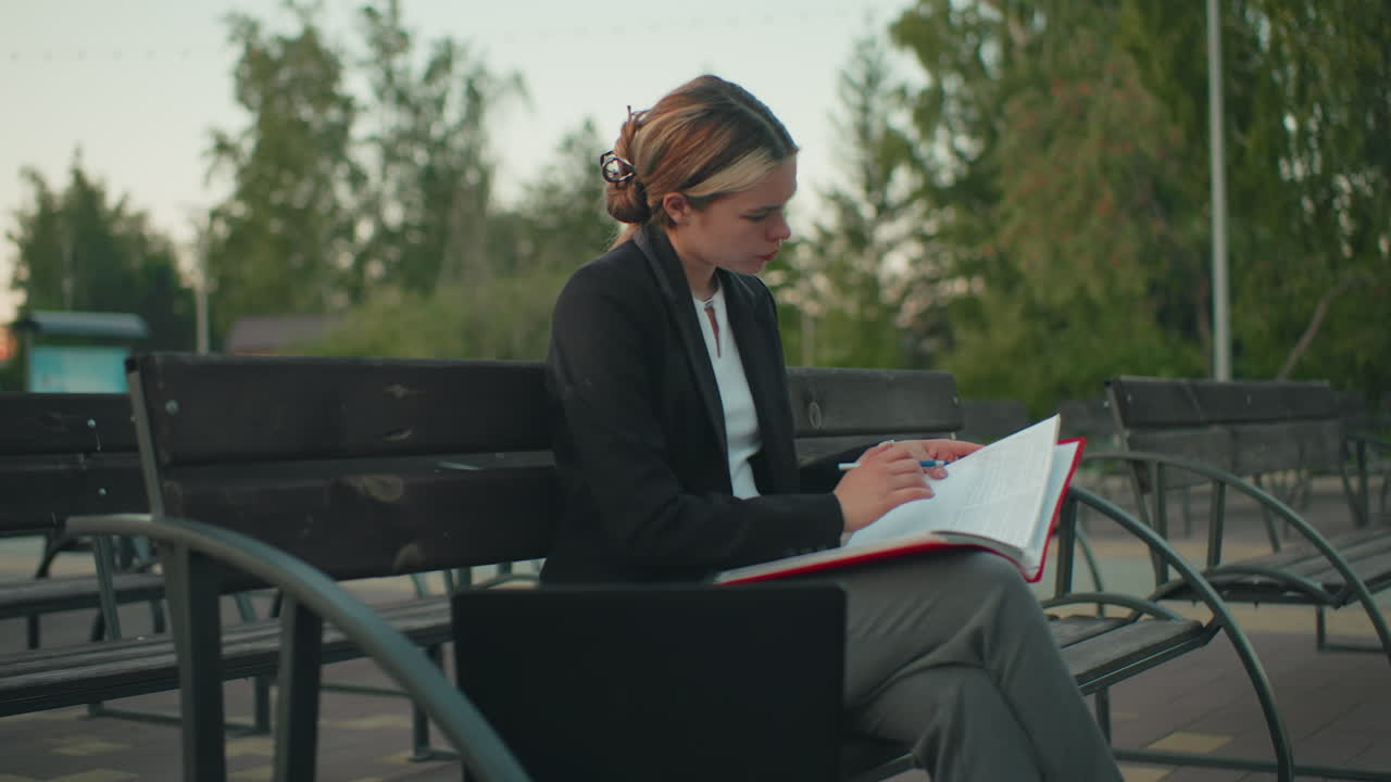 Sidevisning av ung jente som sitter utendørs, dypt fokusert på boken sin mens hun holder en åpen mappe med en penn i hånden, med en laptop og kaffekopp i nærheten i et fredelig bymiljø med bygning og trær bak