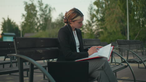 side view young girl seated outdoor deeply focused on her book while holding open folder with pen in hand, laptop and coffee cup nearby in peaceful urban environment with building and trees behind