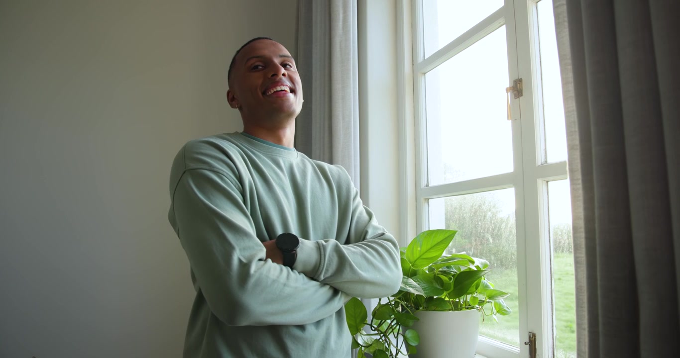 Afroamerikanischer Mann am Fenster mit Pflanze, blickt nach draußen, verschränkt die Arme und lächelt in die Kamera.