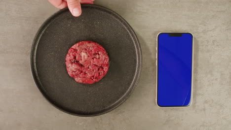 raw hamburger patty on a plate, next to a smartphone