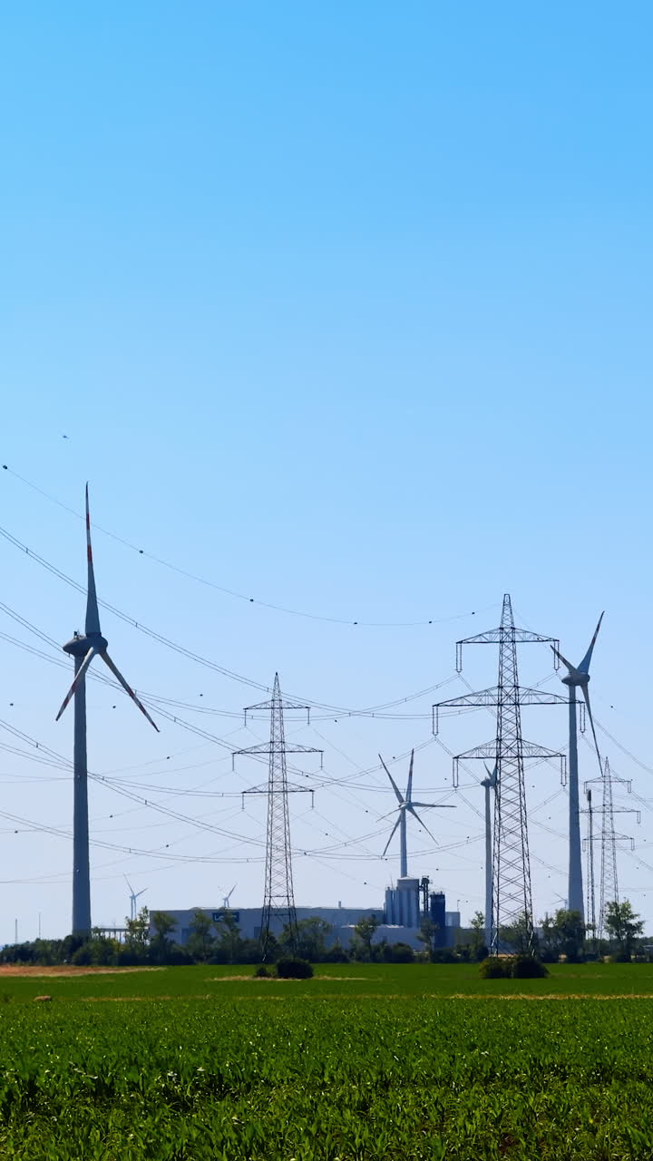 Turbines and lines on clear day. Wind turbines rotate in the breeze while power lines stretch across the bright blue sky and green fields below