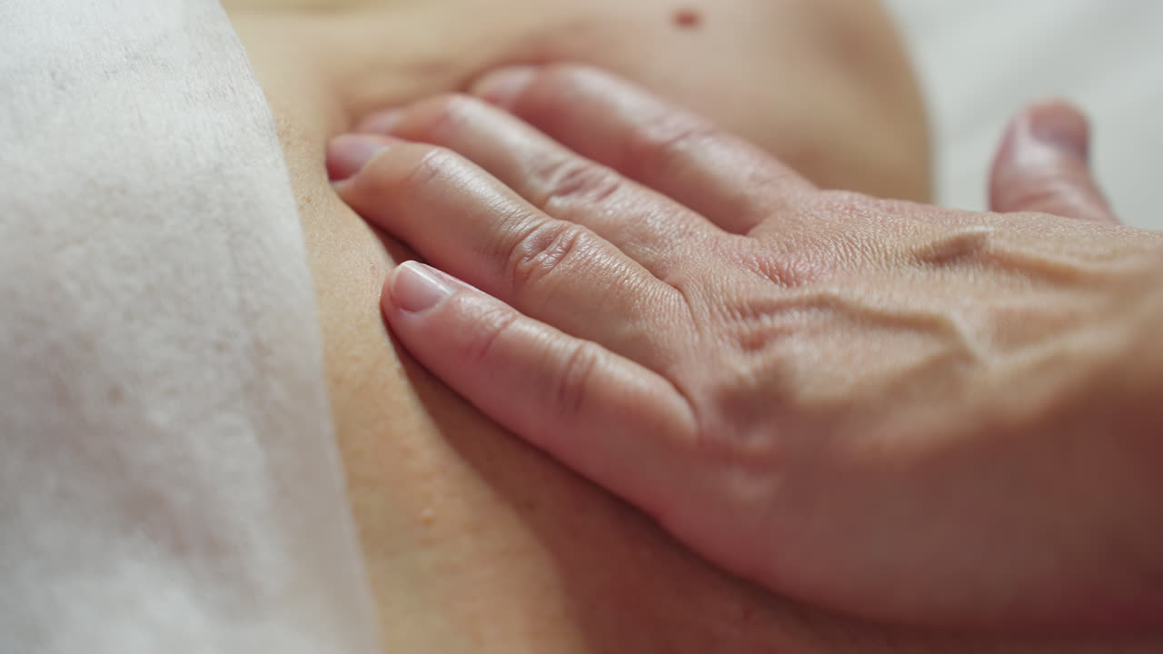Beauty technician glides fingertips through client skin in gentle circular massage motions along chest and neck during relaxing spa session under soft clinical lighting with overhead lamp
