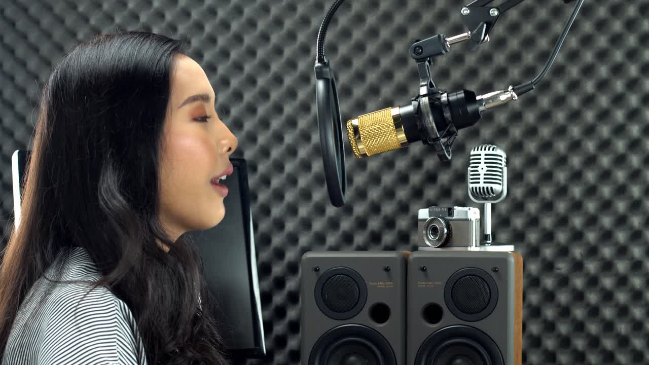 Mulher adolescente asiática com cabelos longos e pretos presos, cantando alto com voz potente sobre microfone condensador suspenso e equipamento.