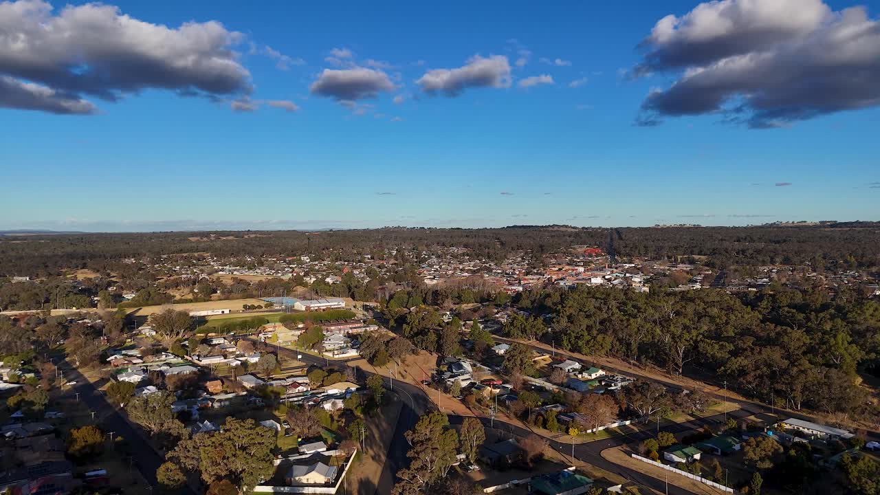 Drone camera glides above a small Australian town, revealing new residential areas, natural woodland, and street layout under clear blue sky with afternoon sunlight