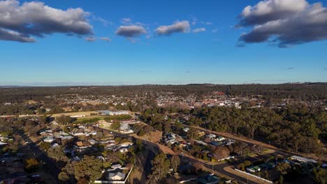 drone camera glides above a small australian town, revealing new residential areas, natural woodland, and street layout under clear blue sky with afternoon sunlight