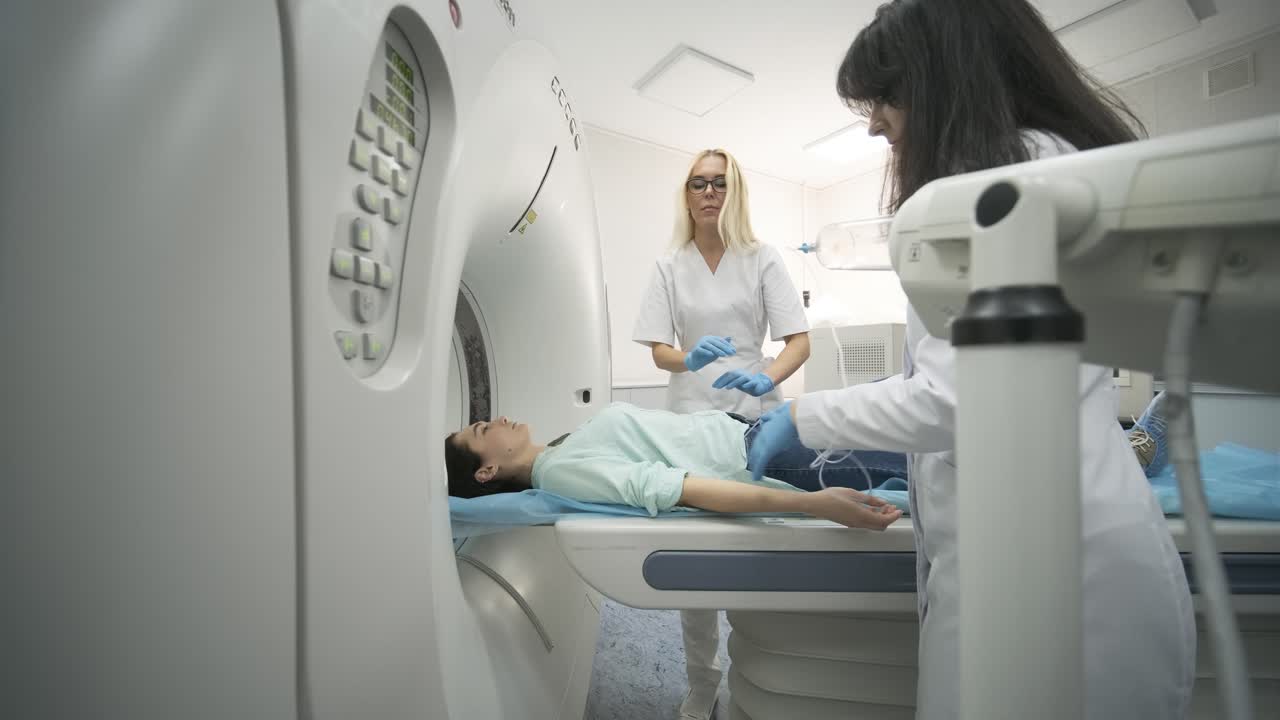 Two radiologists inserts an intravenous catheter to inject contrast to female patient who is undergoing CT or MRI scan in modern medical clinic. Patient lying on a CT or MRI scan bed, moving inside the machine.